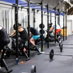 Row of people riding workout machinery bikes in fitness gym and trainer assists nearby.